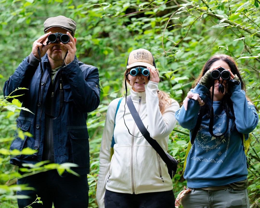 Photo features three people, the two on either side are looking up and to the left through binoculars. The one in the middle is wearing sunglasses and waving and the viewer.