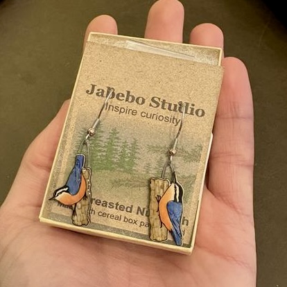 Photo features a set of asymmetrical bird earrings held in the palm of a hand.