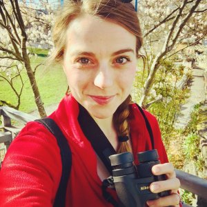Photo features Dr. Olivia Sanderfoot, standing outside wearing red and holding a pair of binoculars.