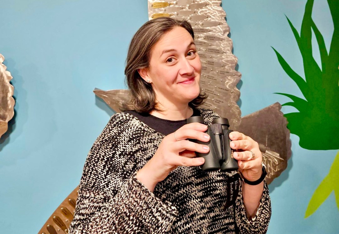 Photo features a woman in a grey sweater holding a pair of small binoculars and smiling.