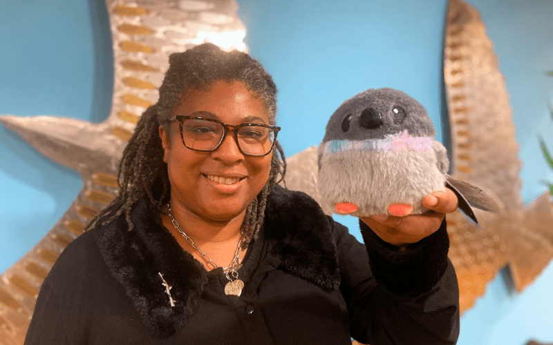 Photo features a woman wearing a black top and glasses holding a small stuffed pigeon plush toy.