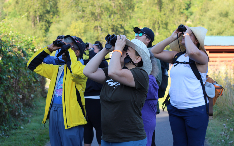 Photo features a group of people standing together and looking up through binoculars.