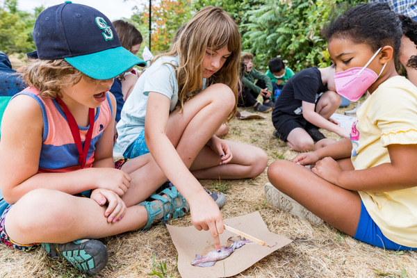 Three children sit cross-legged on the ground examining a squid specimen.