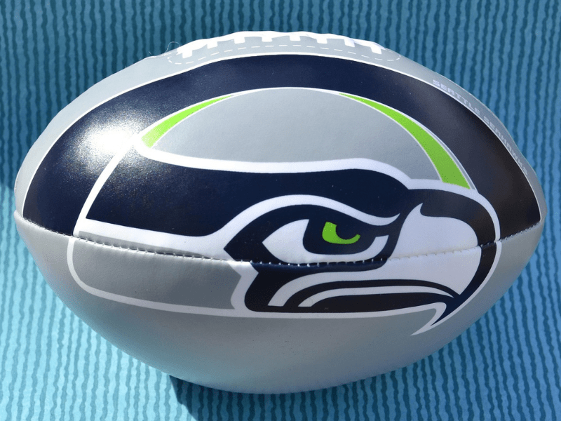 Photo features a gray football with the blue, white, gray, and green logo of the Seattle Seahawks. Background is blue.