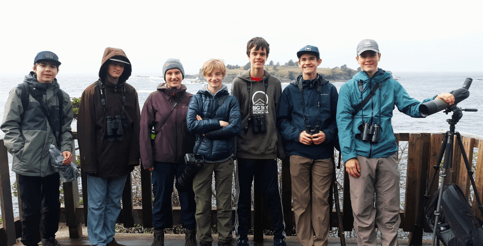 Photo features a Sandhill Crane, a large bird, standing on one leg with wings open.
